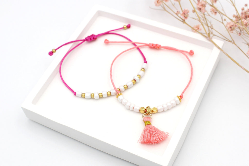 Sommerarmband bunt im 2er Set für Damen, Pink und Korallen farbenes Makrameeband mit weiß und goldenen Details und geflochtenem Makramee Verschluss auf weißem Tablett liegend